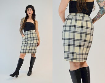 May include: A cream-coloured pencil skirt featuring a black and blue plaid pattern. The skirt has a high waist, a straight cut, and a small pocket on the front. The skirt is worn with black boots.