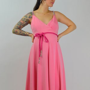 Może przedstawiać: Różowa sukienka z dekoltem w serek i cienkimi ramiączkami ozdobionymi błyszczącymi detalami. Sukienka posiada pas w talii z różowej liny i zwiewną spódnicę. Stylowy, elegancki strój.