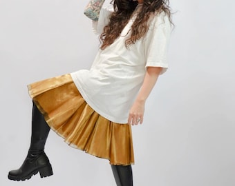 May include: A person wearing a white oversized t-shirt and a gold skirt with black boots. The skirt has a layered design. The boots are knee-high with a chunky heel. The person has long, dark hair and tattoos on their arm.