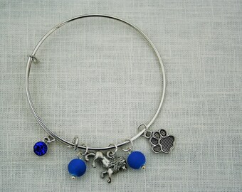 May include: A silver bangle bracelet with blue charms, including a paw print, a blue crystal, and two blue glass beads. The bracelet also features a silver charm of a running horse.