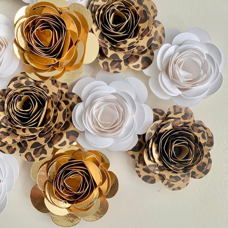 Op de afbeelding: Een assortiment van papieren bloemen in verschillende kleuren en patronen. De bloemen zijn gemaakt van papier en hebben een gelaagd, 3D-effect. De bloemen zijn in verschillende kleuren, waaronder wit, goud en bruin. De bloemen zijn in een cirkelvormige opstelling gerangschikt.