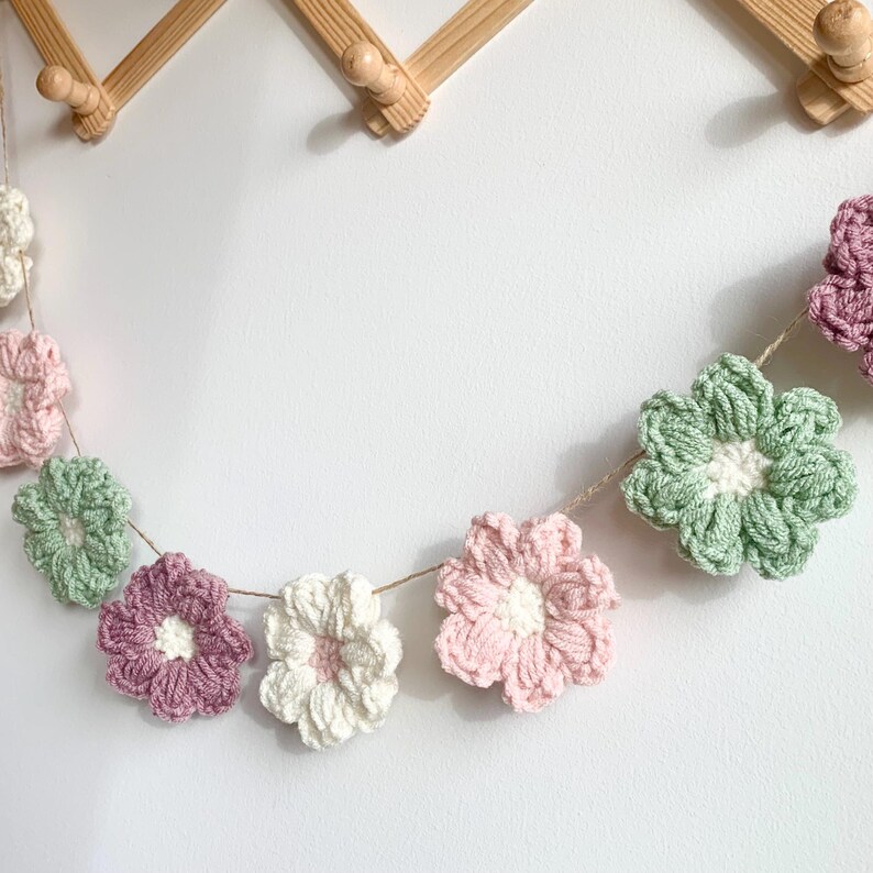 May include: A garland of crocheted flowers in shades of pink, green, and white. The flowers are strung together with twine and hang from a wooden wall.
