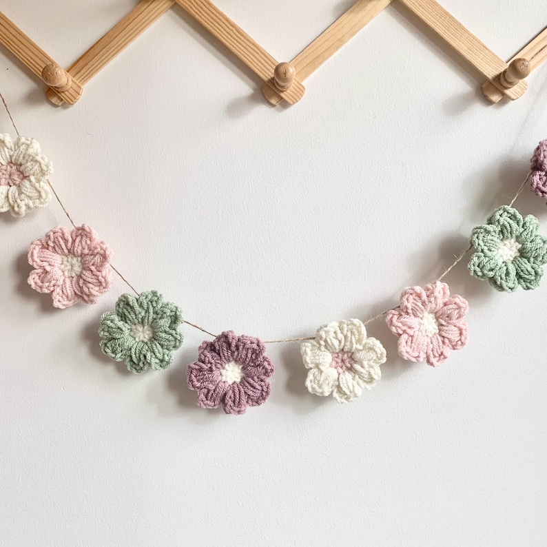 May include: A garland of crocheted flowers in shades of pink, green, and white. The flowers are strung together with twine and hang from a wooden wall rack.