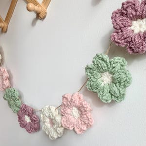 May include: A garland of crocheted flowers in pink, green, and white. The flowers are strung together with twine and hang against a white wall.