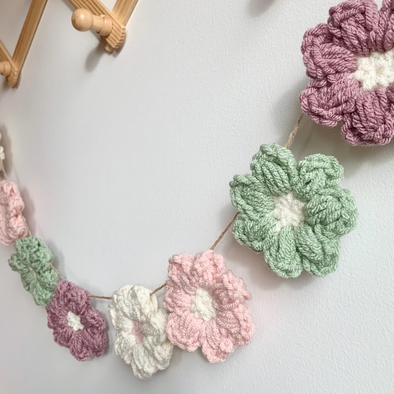 May include: A garland of crocheted flowers in pink, green, and white. The flowers are strung together with twine and hang against a white wall.