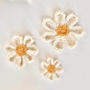 May include: Three crocheted daisies in various sizes, with white petals and yellow centers, on a white background.