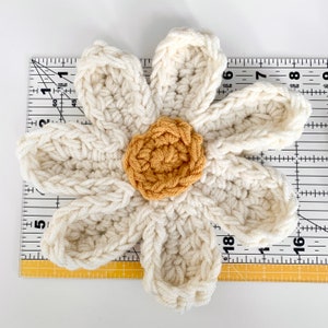 May include: A crocheted white daisy flower with a yellow center. The flower is about 6 inches in diameter and is laid on top of a ruler.