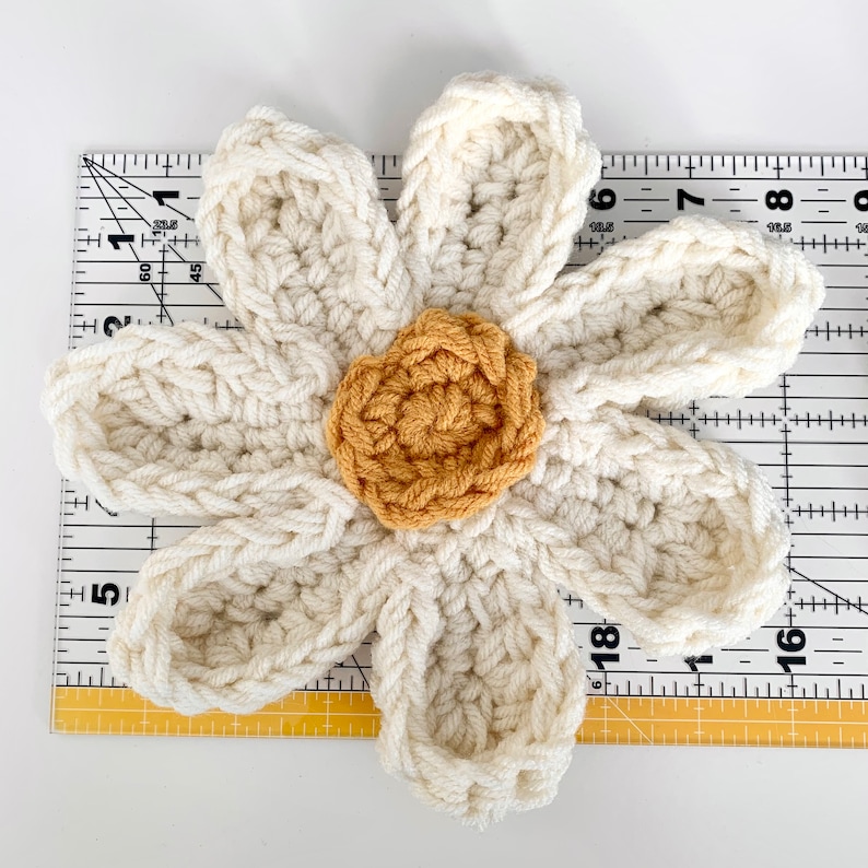May include: A crocheted white daisy flower with a yellow center. The flower is about 6 inches in diameter and is laid on top of a ruler.