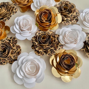 Op de afbeelding: Een verzameling papieren bloemen in verschillende kleuren en patronen. De bloemen zijn gemaakt van papier en hebben een gelaagde, gerimpelde uitstraling. Sommige bloemen zijn wit, andere zijn goud en weer andere zijn bruin met een luipaardprint.
