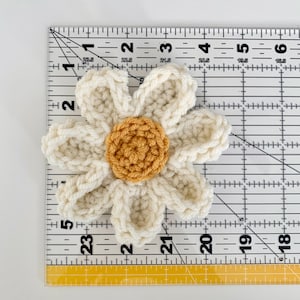 May include: A crocheted white daisy flower with a yellow center. The flower measures approximately 2 inches in diameter.