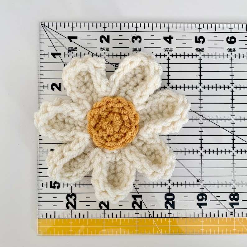 May include: A crocheted white daisy flower with a yellow center. The flower measures approximately 2 inches in diameter.