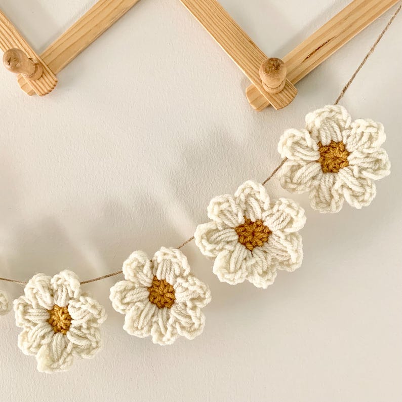 May include: A garland of crocheted white daisies with yellow centers hangs from a wooden rack. The daisies are strung together with twine.