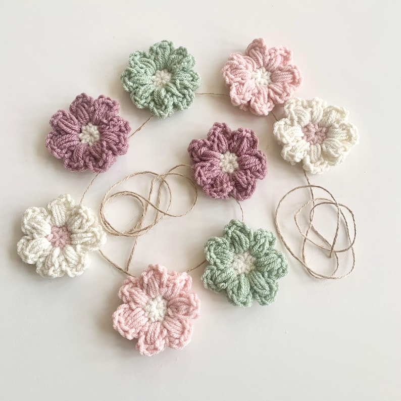 May include: A garland of crocheted flowers in shades of pink, green, and white. The flowers are strung together with twine and are perfect for adding a touch of whimsy to any space.