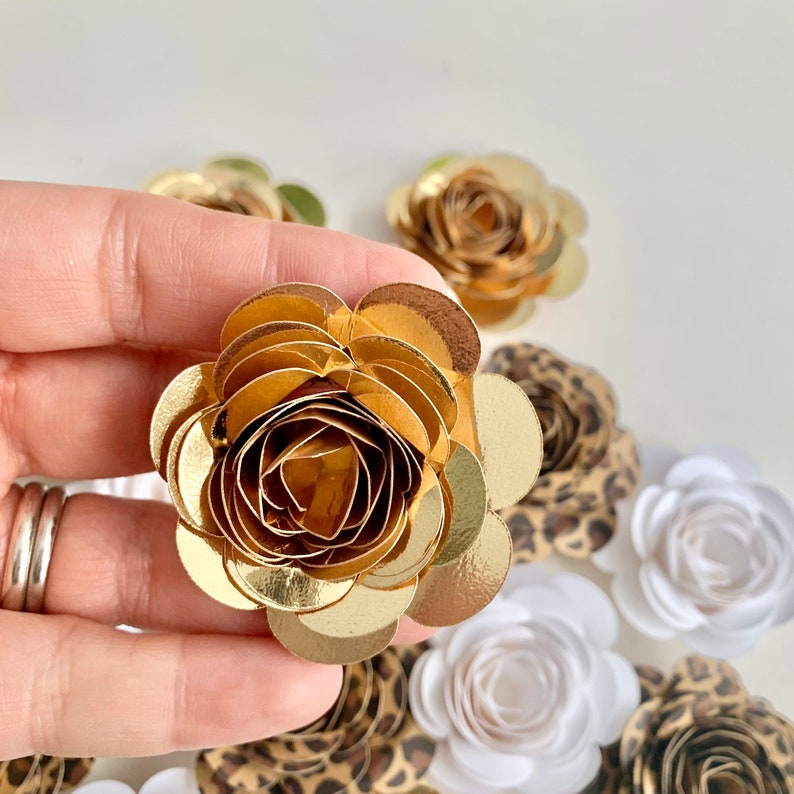 Op de afbeelding: Een gouden papieren bloem met een gelaagd rosetontwerp. De bloem wordt omringd door andere papieren bloemen in verschillende kleuren en patronen.