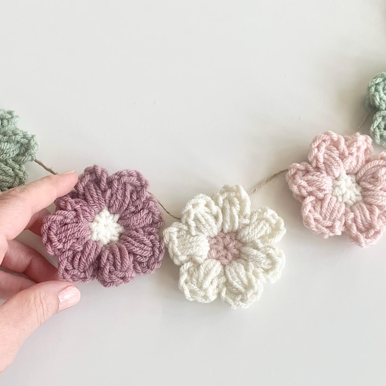 May include: A garland of crocheted flowers in shades of pink, white, and green. The flowers are connected by a string and are arranged in a row.