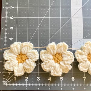 May include: A garland of crocheted white daisies with yellow centers. The daisies are strung together with twine and are displayed on a gray cutting mat.
