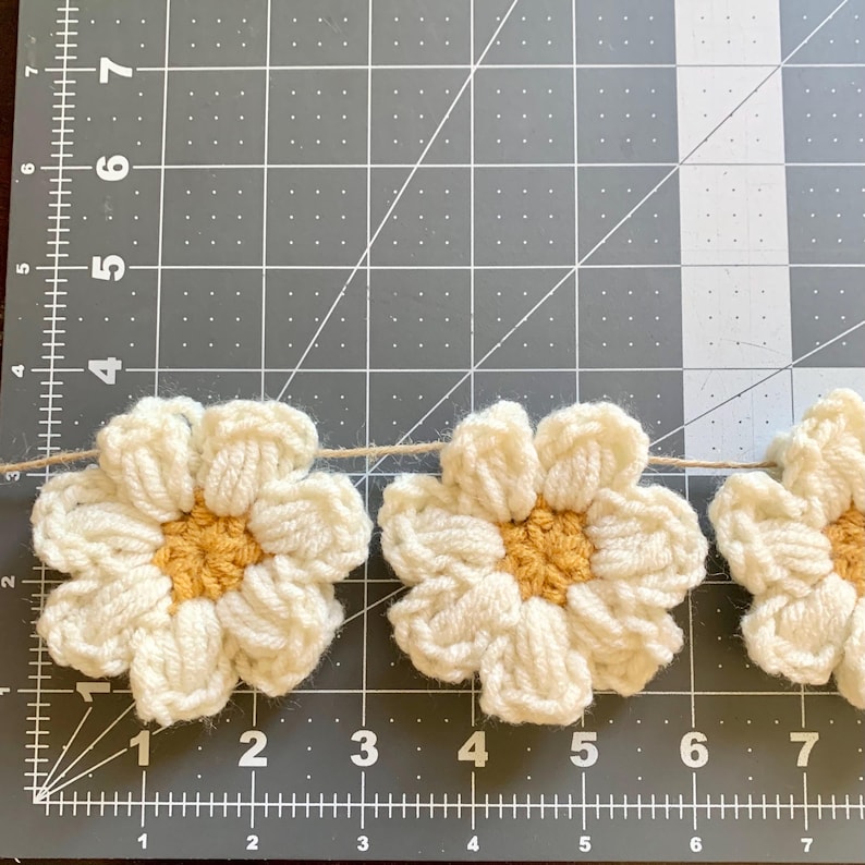 May include: A garland of crocheted white daisies with yellow centers. The daisies are strung together with twine and are displayed on a gray cutting mat.