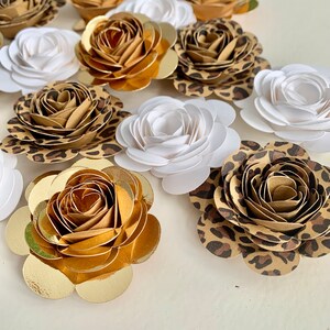 Op de afbeelding: Een verzameling papieren bloemen in verschillende kleuren en patronen. De bloemen zijn gemaakt van papier en hebben een gelaagde, gerimpelde uitstraling. Sommige bloemen zijn wit, terwijl andere bruin zijn met een luipaardprint. De bloemen zijn willekeurig gerangschikt op een wit oppervlak.