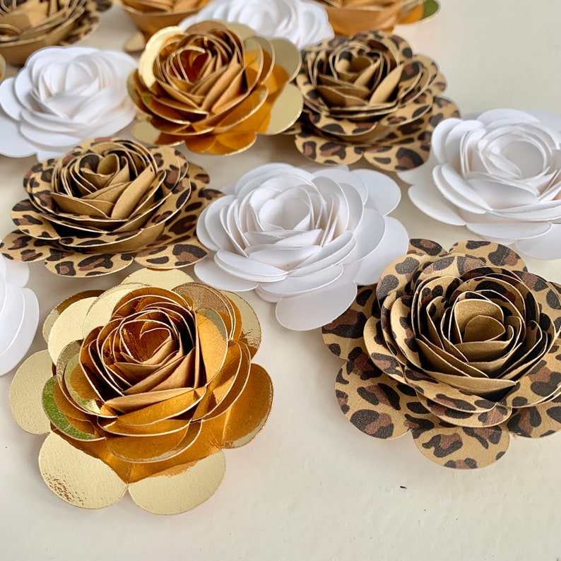 Op de afbeelding: Een verzameling papieren bloemen in verschillende kleuren en patronen. De bloemen zijn gemaakt van papier en hebben een gelaagde, gerimpelde uitstraling. Sommige bloemen zijn wit, terwijl andere bruin zijn met een luipaardprint. De bloemen zijn willekeurig gerangschikt op een wit oppervlak.