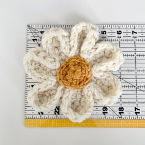 May include: A crocheted white flower with a yellow center. The flower is approximately 6 inches in diameter and is measured against a ruler.