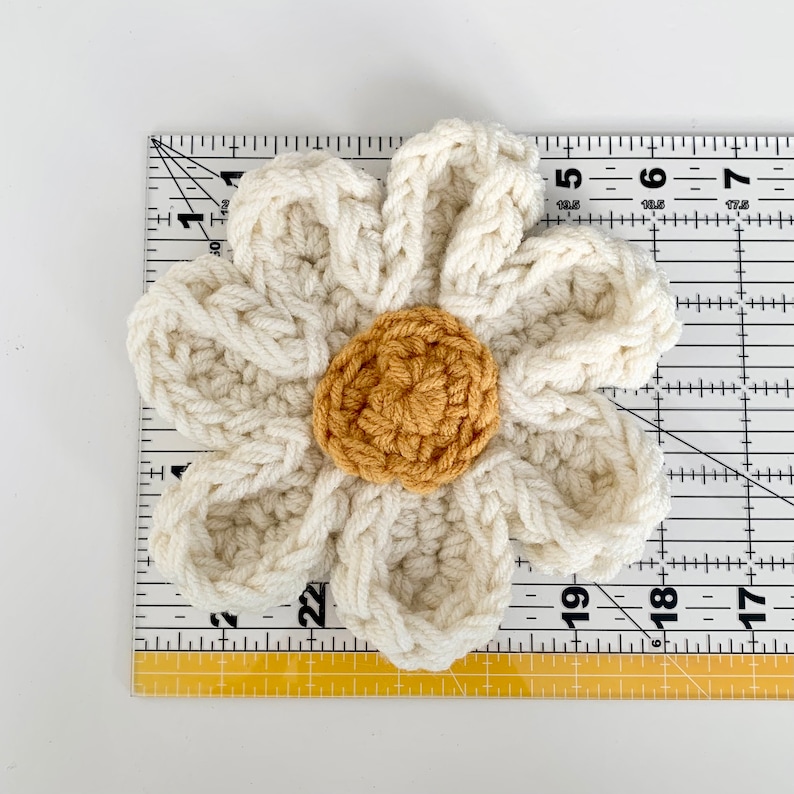 May include: A crocheted white flower with a yellow center. The flower is approximately 6 inches in diameter and is measured against a ruler.