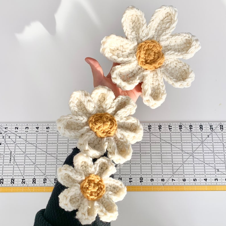 May include: Three crocheted white daisy flowers with yellow centers. The flowers are arranged in a row, with the largest flower at the top and the smallest flower at the bottom.
