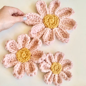 May include: Three crocheted pink flowers with yellow centers. The flowers are different sizes and are arranged on a white surface.