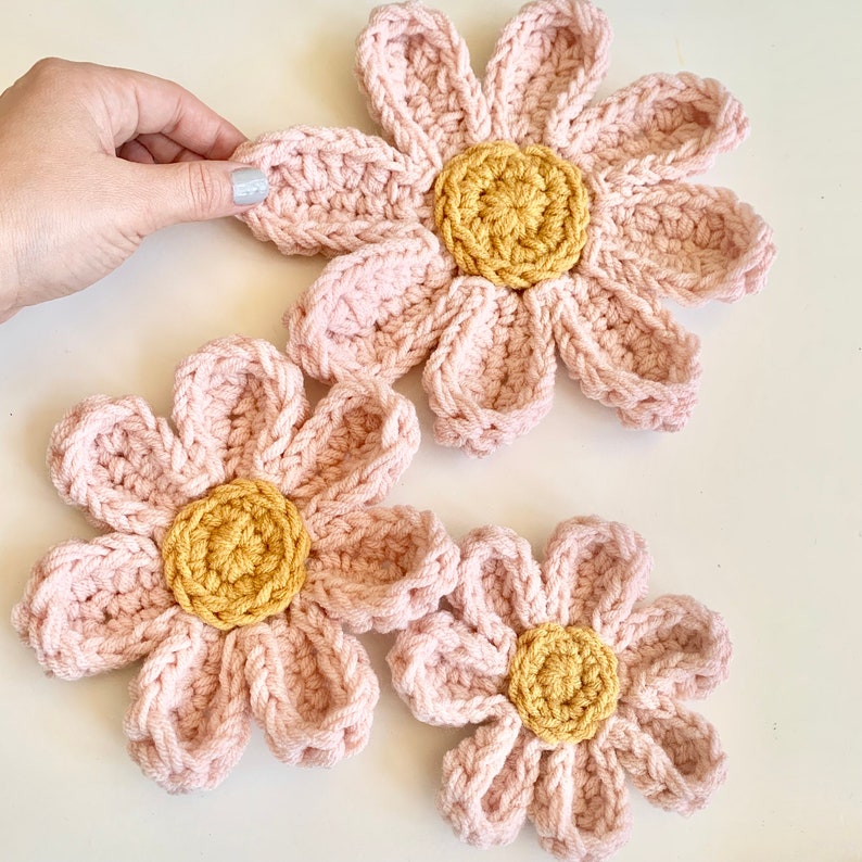 May include: Three crocheted pink flowers with yellow centers. The flowers are different sizes and are arranged on a white surface.