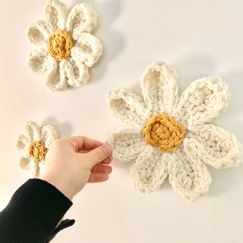 May include: Three crocheted white daisies with yellow centers. The daisies are hanging on a white wall.