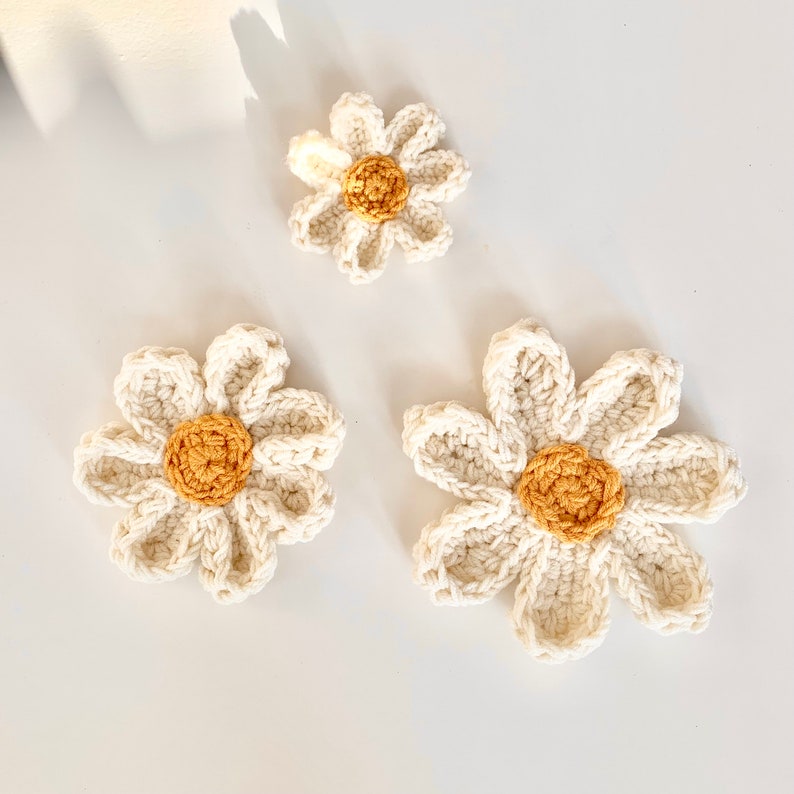 May include: Three crocheted daisy flowers in various sizes. The flowers are white with yellow centers.