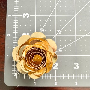 Op de afbeelding: Een gouden papieren bloem met meerdere lagen bloemblaadjes. De bloem is ongeveer 5 cm in diameter.