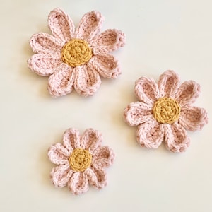 May include: Three crocheted pink flowers with yellow centers. The flowers are different sizes and are arranged on a white background.