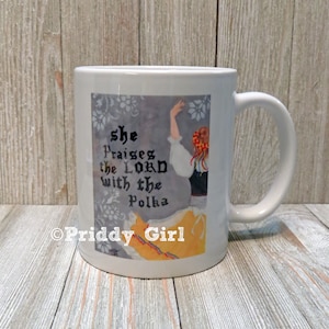 May include: White ceramic mug featuring a design with the text "She Praises the Lord with the Polka" and an illustration of a woman in traditional attire. The mug has a curved handle and a glossy finish.