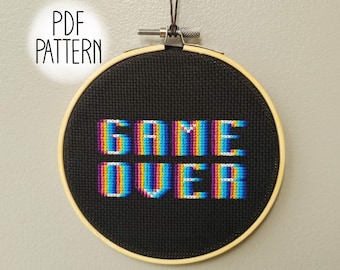 May include: A black embroidery hoop with a colorful pixelated cross-stitch design that reads "GAME OVER".