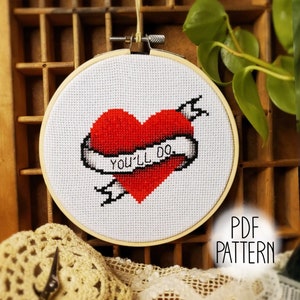 May include: A cross-stitch pattern featuring a red heart with an arrow through it and a banner that reads "You'll Do". The pattern is displayed in a wooden embroidery hoop.