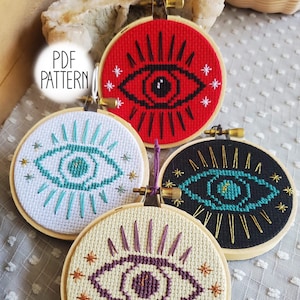 May include: Four embroidery hoops with different colored backgrounds and a stylized eye design with radiating lines. The eye designs are in red, teal, purple, and turquoise. The text "PDF PATTERN" is visible in the top left corner of the image.