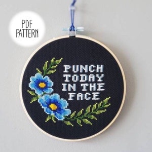 May include: A black embroidery hoop with a cross-stitch design featuring blue flowers and green leaves. The text "Punch Today In The Face" is stitched in white on a black background.