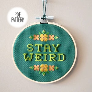 Puede incluir: Un aro de bordado verde con la frase "STAY WEIRD" bordada en hilo amarillo. El aro está decorado con pequeñas flores naranjas y amarillas.