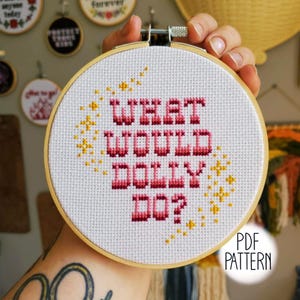 May include: A circular wooden embroidery hoop containing white Aida fabric with a cross-stitch design. The text reads "WHAT WOULD DOLLY DO?" in pink and red gradient lettering, surrounded by small yellow cross-stitched stars and sparkles.