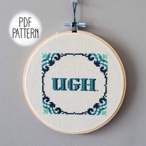 May include: A white embroidery hoop with a blue and green cross-stitch design. The design features a decorative frame with the word "UGH" in the center.