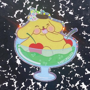 May include: A sticker featuring a yellow cartoon character in a sundae glass with cherries. The character has a brown beret and is eating a sweet treat. The sundae has green liquid and the text "BABYLIPGLOSS".