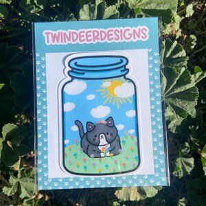 May include: A sticker featuring a black and white cat with a flower crown inside a blue mason jar with a sun and clouds. The jar is on a blue background with white paw prints.
