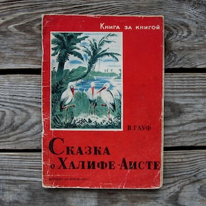May include: A red book cover with the title "Сказка о Халифе-Аисте" in black text. The book cover features an illustration of three white storks standing in a green field with a palm tree in the background. The book cover also includes the text "Книга за книгой" and "В. Гауф".