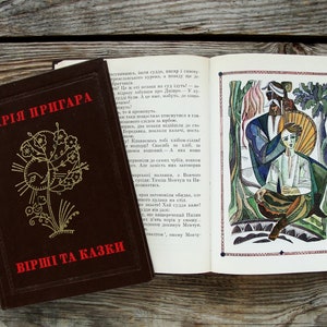 Puede incluir: Un libro marrón cerrado con el título "MARIYA PRYHARa" y el texto "VIRSHI TA KAZKI" en la portada. El libro está abierto en una página con una ilustración de dos personas en un entorno de bosque. La ilustración está en un estilo de arte colorido y estilizado.