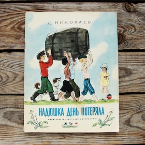 May include: A vintage children's book cover with a colorful illustration of four children carrying a large wooden barrel. The book title is "Nadioska Den Potierala" and the author is "D. Nikolaev".