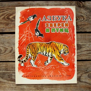 Op de afbeelding: Vintage kinderboekcover met een tijger op een oranje achtergrond. Twee zwart-witte vogels zijn ook afgebeeld. De titel is in het Russisch. Het boek komt uit 1987.