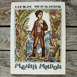 Puede incluir: Una portada de libro vintage con una ilustración colorida de un niño pequeño de pie en un campo de flores y plantas. El título del libro es "Мамий Мирон" y el autor es "Іван Франко".