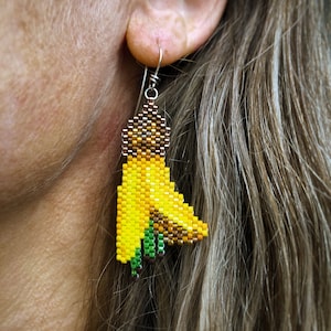 Boucles d'oreilles fleur kōwhai, tissées avec des perles de rocaille Miyuki Delica, sur des crochets de boucle d'oreille français en argentium 93,5 dans une boîte en papier kraft.