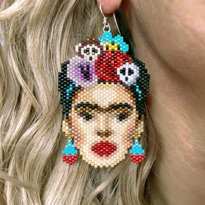 Puede incluir: Pendientes hechos a mano con cuentas que representan a Frida Kahlo. Los pendientes están hechos con pequeñas cuentas de colores, que representan sus rasgos icónicos, incluyendo una ceja unida, labios rojos y un tocado floral. Tienen un gancho plateado.