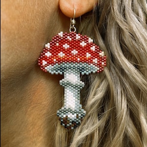 May include: A mushroom-shaped earring made of beads. The cap is red with white spots, the stem is grey and white. The earring hangs from a silver hook. A unique piece of jewellery.
